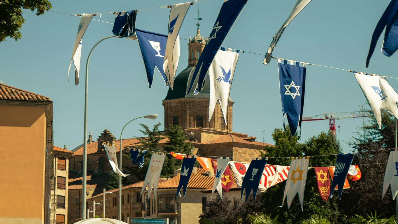 Colorful medieval-style flags with Jewish symbols and doves hanging in the streets of Salamanca, Spain, during a cultural or interfaith festival, with a historic dome and stone buildings in the background.