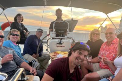 A group of travelers aboard a sailboat during sunset in Lisbon, near Belém Tower, with glasses of wine and traditional Portuguese snacks.