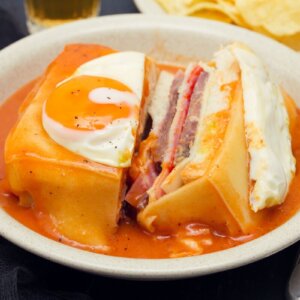 Traditional Francesinha sandwich from Porto, covered in melted cheese and a rich sauce, with a fried egg on top.