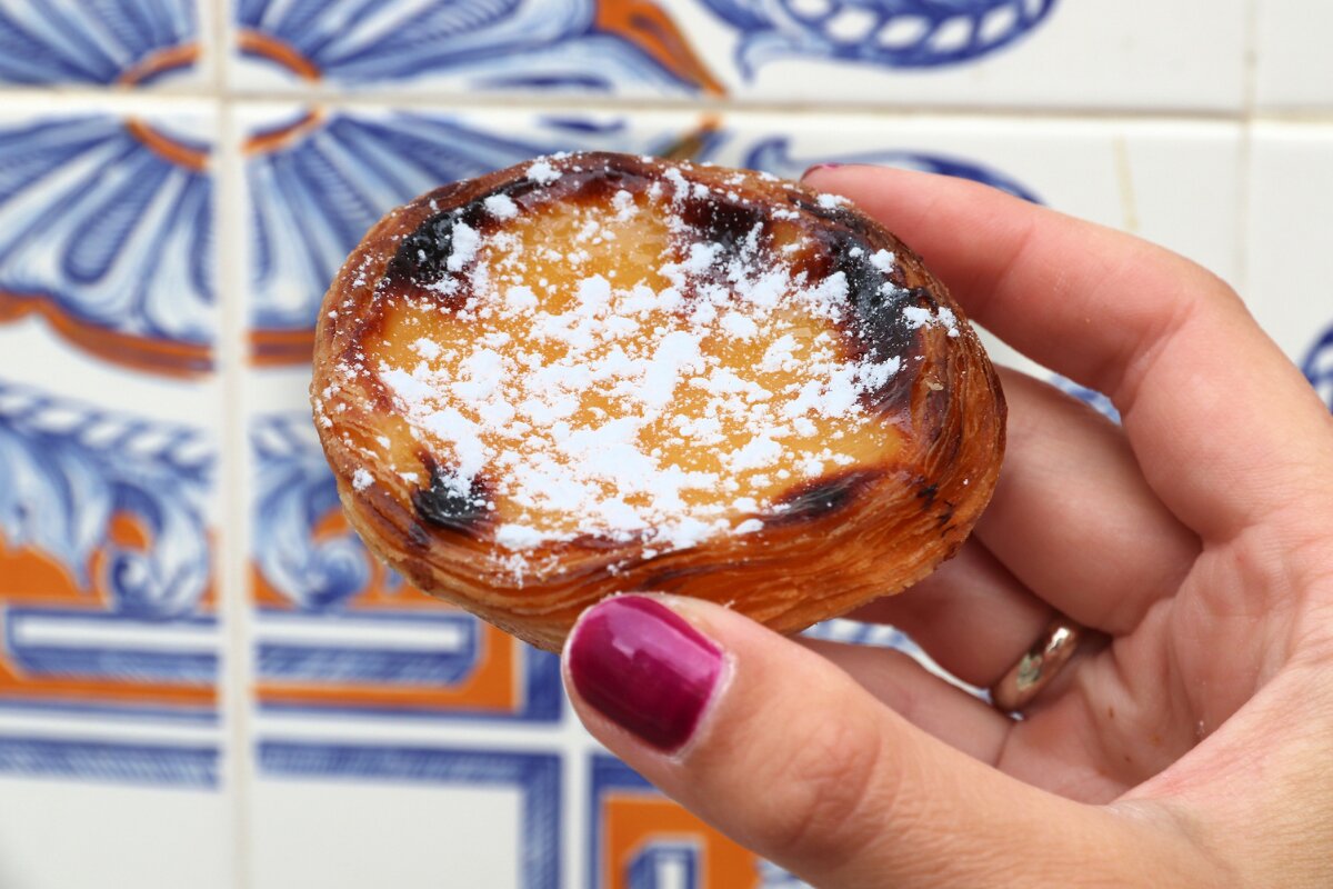 Traditional Portuguese pastel de nata dusted with powdered sugar, held in front of classic blue and white azulejo tiles.